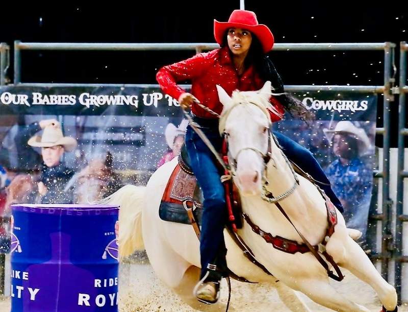 African American teen cowgirl racing around a blue barrel on a white horse. 