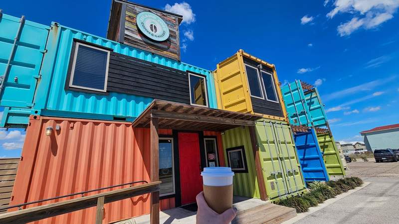 Holding a to-go coffee cup in front of colorful shipping containers that were repurposed for a coffee shop 