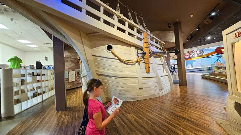 A young girl walking around a museum with a mock wooden ship