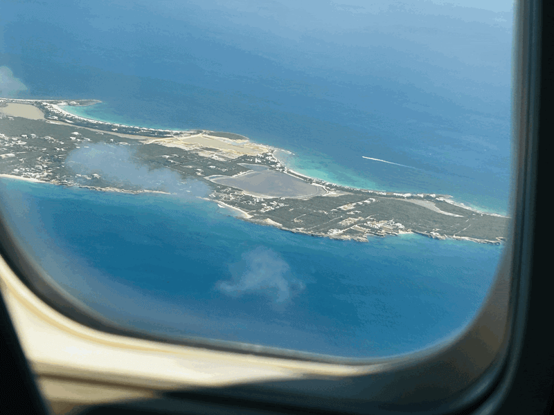 View from an airplane window looking down at the lush green hills, turquoise ocean waters, and coastline of St. Maarten during descent.