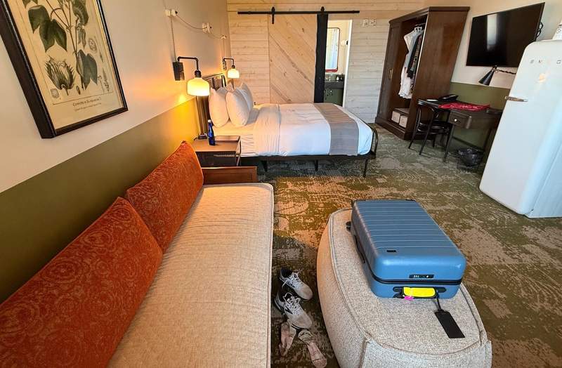 upholstered coffee table doubles as suitcase holder in front of sofa near hotel queen bed. Refrigerator stands next to writing table and armoire near sliding barn door opening to bathroom.
