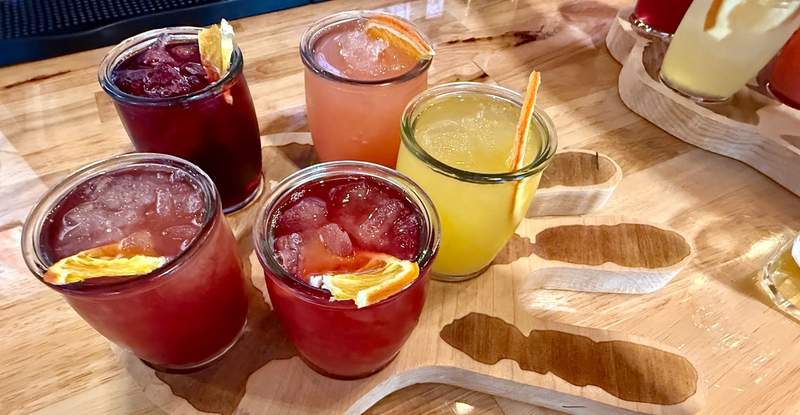Five glasses filled with different colors of sangria stand on a wooden tray shaped like an open hand with the palm up. One glass holds a straw and four have slices of orange.