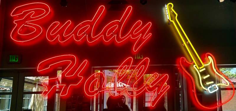 Red neon letters spell the Buddy Holly name on a sign reflecting into windows. Neon of red and yellow creates the shape of a guitar.