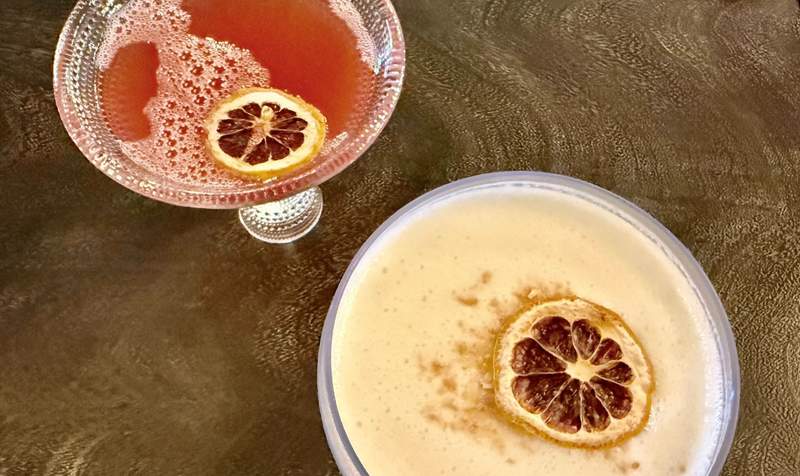 Two round glasses with short stems hold Chilton cocktails. One is cream color and the other like red grapefruit. A lemon slice floats in each.