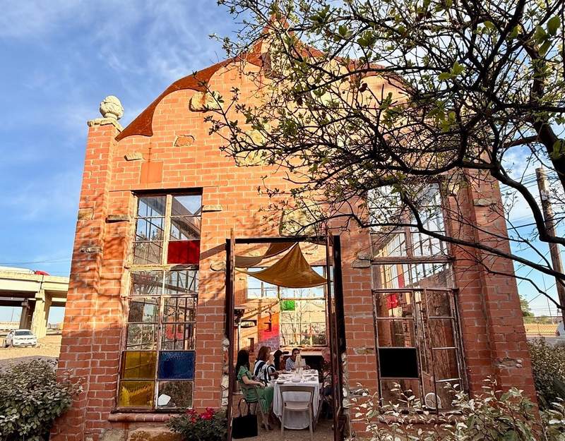 Blue sky frames the pale brick greenhouse with long tall window panes and a wide open door. Tables fill the inside for fine dining instead of greenhouse plants.