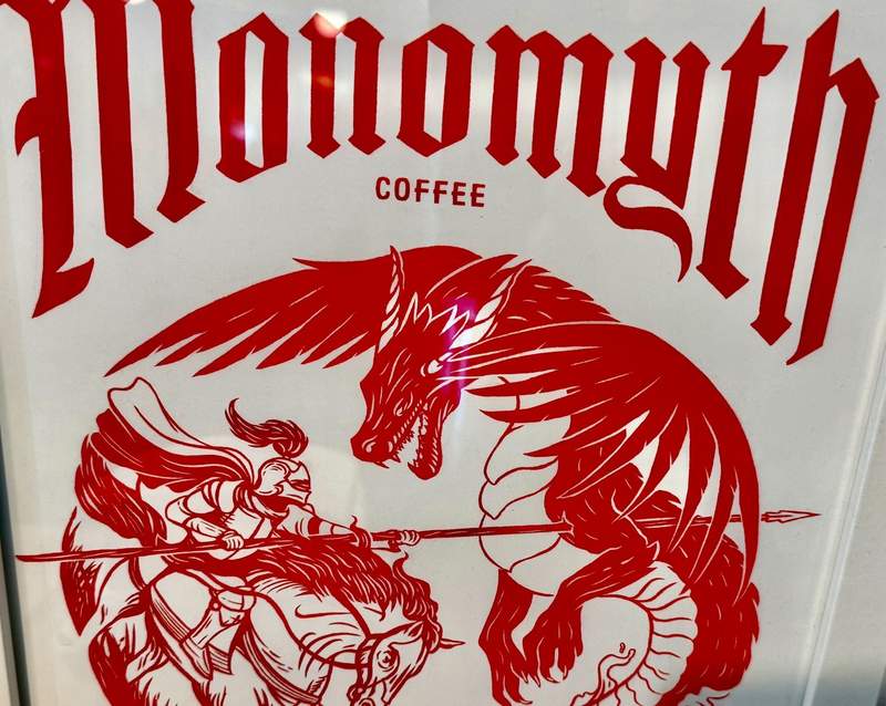 White poster with red ink declares Monomyth Coffee on the top. A red and white dragon menaces a warrior with a long spear on horseback.
