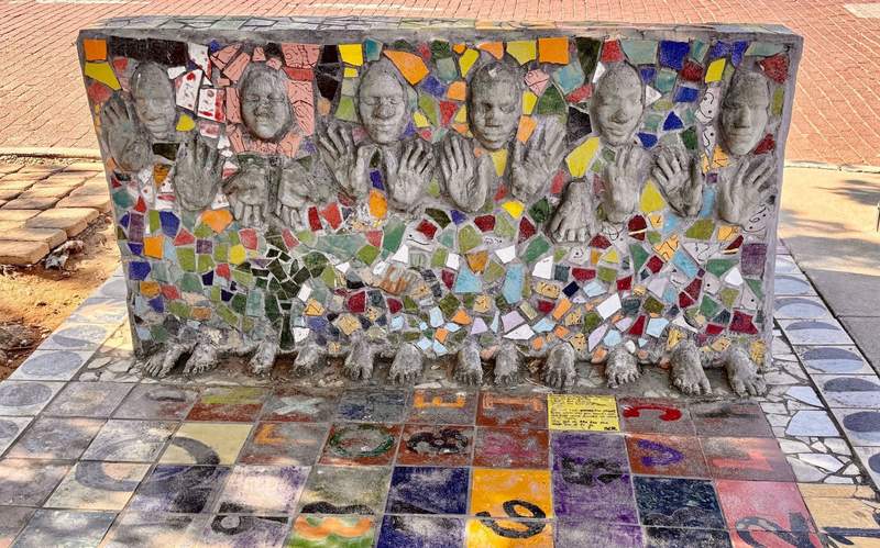 A wall of hands and feet and faces with closed eyes is surrounded with mosaic pieces of glass in shapes and colors to form back of a sidewalk bench.