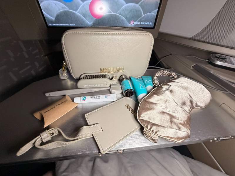 Airline tray table with X MOSHINO travel kit spread out with toothbrush, toothpaste, luggage tag, eye mask, earplugs and personal care items.