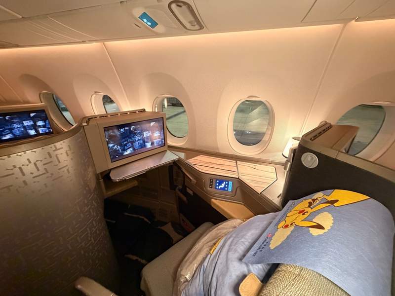 Business class seat on China Airlines features neutral colors, a lie-flat seat, a Pikachu headrest cover, a Pikachu pillow, a lamp and views through the porthole window.