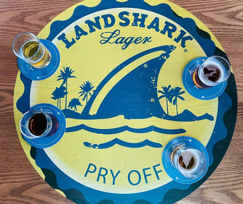 A flight of four diverse craft beers in small tasting glasses, sitting on a branded LandShark wooden paddle board.