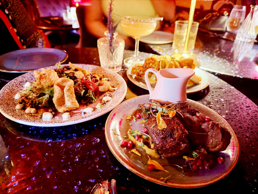 Craft cocktails and upscale shareable plates, including lamb chops and beef carpaccio, on a dark table inside a moody, intimate speakeasy lounge