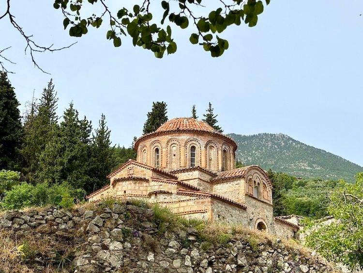 An exterior view of a medieval Byzantine church in Mystras, Greece, showing historic stone walls, an octagonal tiered structure, and a weathered red-tiled roof.