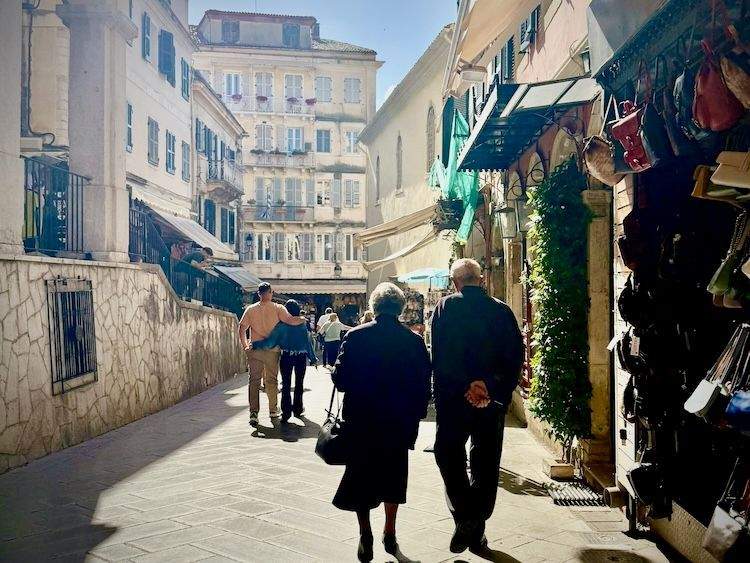 An older couple walking side-by-side down a narrow, sunlit street in Corfu Old Town, flanked by tall Venetian-style historic buildings.