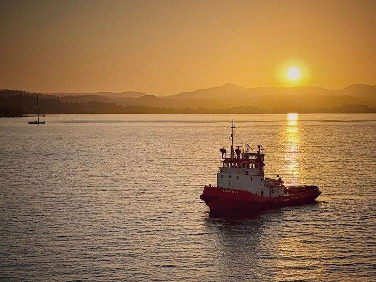 A vibrant red and white tugboat navigating the calm sea at sunset, guiding a larger ship away from the shores of Corfu Island.