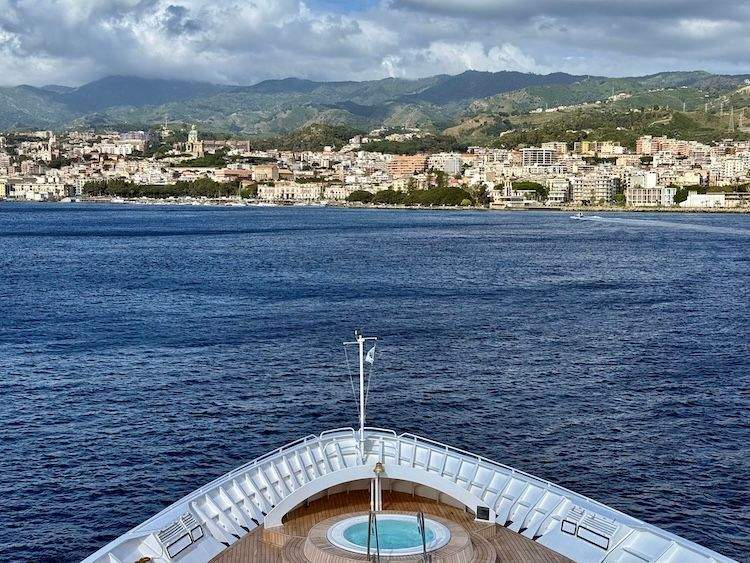 The pointed bow of the Star Legend cruise ship slicing through calm, glassy blue waters as it approaches the coast of Messina, Sicily in the morning light.