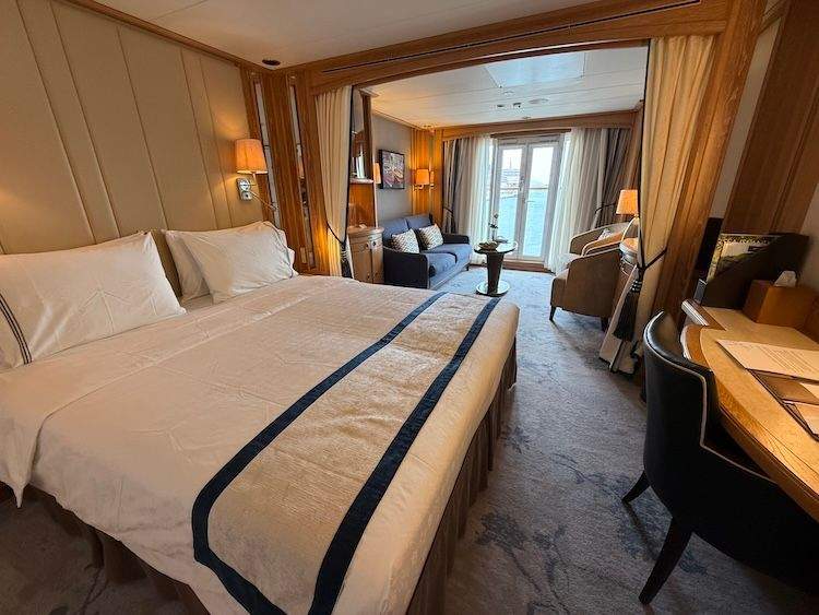 A bright, all-suite luxury cabin aboard the Star Legend cruise ship, featuring a neatly made bed, a small desk, a comfortable sofa and chairs, and a glass door opening to a balcony view.