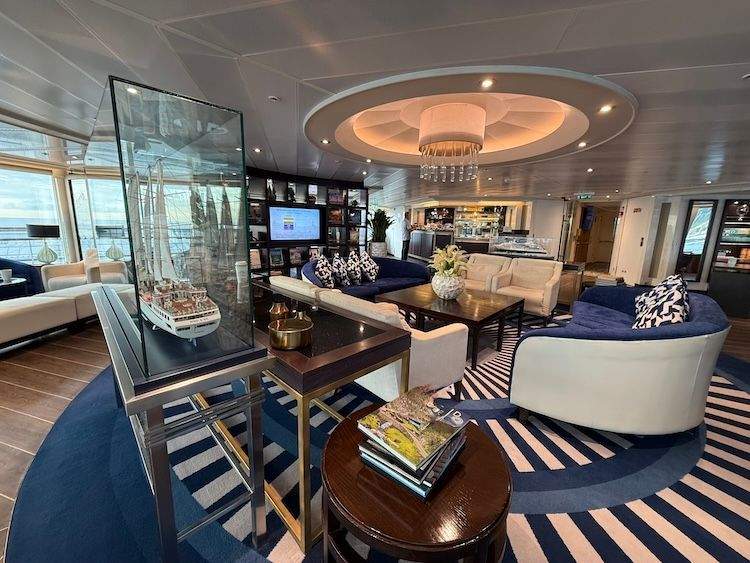 The interior of the Yacht Club lounge on a ship, featuring a blue and white striped rug, comfortable modern seating areas, a coffee bar, and large windows with panoramic sea views.