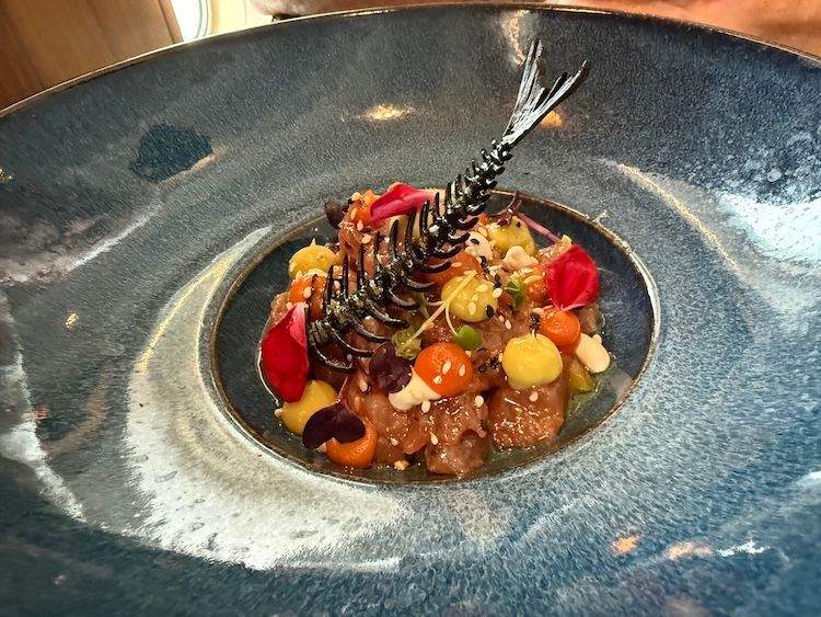 An elegant tuna tartare appetizer served in a blue bowl, topped with bright, colorful garnishes and an artistic, delicate edible fish-bone decoration.