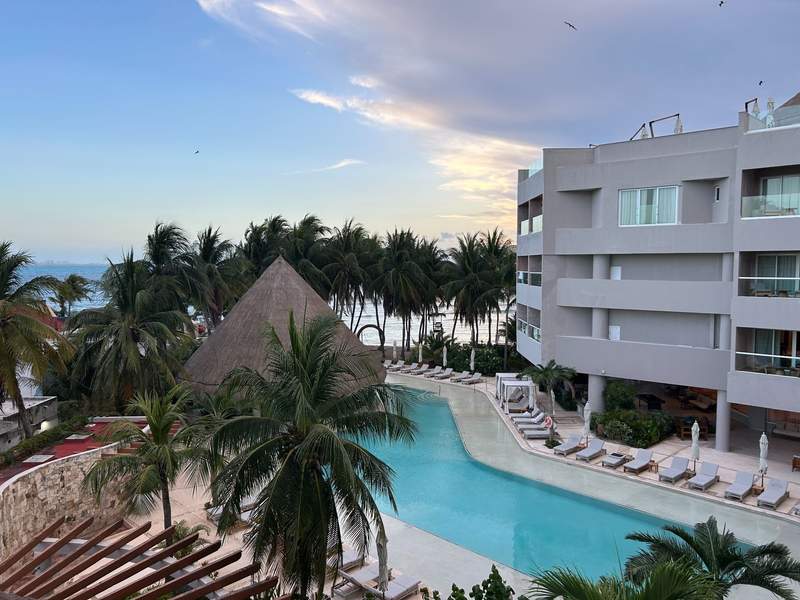 Aerial view of the pool at Almare, a Luxury Collection Resort in Isla Mujeres, surrounded by palm trees, lounge chairs, and a thatched-roof pavilion, with the Caribbean Sea visible in the background.