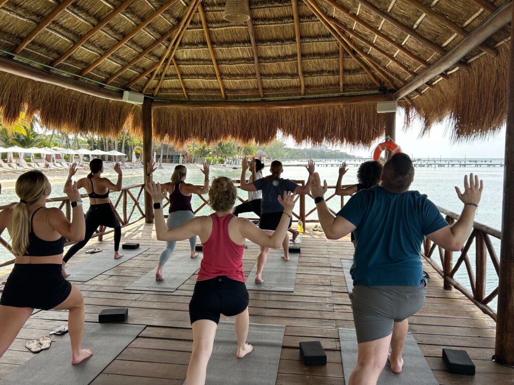 Guests participate in a yoga class on a wooden pavilion with a thatched roof, overlooking turquoise water at Almare, a Luxury Collection Resort, Isla Mujeres