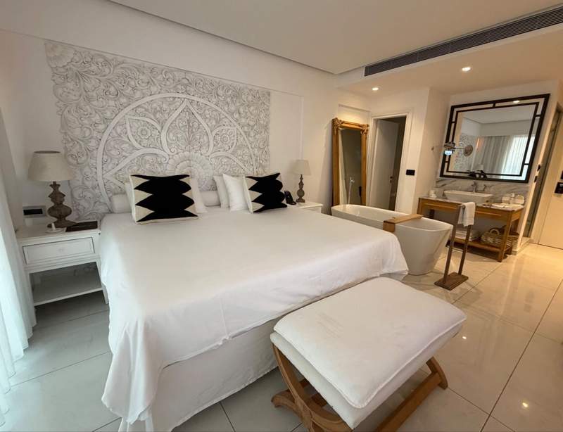 A hotel bedroom features all-white linens, a carved wooden headboard, marble tile floors and a large soaking bathtub.