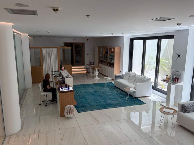 A serene, cool reception area leading to spa rooms. There's a white sofa, white marble tiles, a blue rug, and a woman sitting behind a consultation desk.