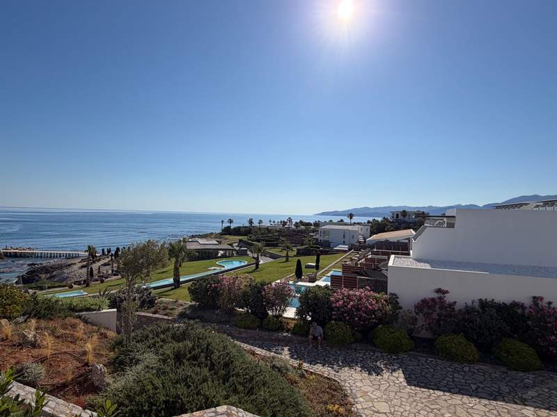 A view of a sweeping beach on the Aegean Sea. White resort suites overlook the sea, there is also a decorative pool, palm trees and other native greenscape.