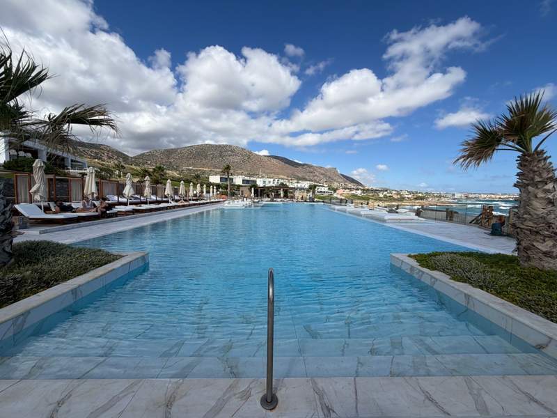 An expansive swimming pool is framed by the Aegean Sea and mountains. There are palm trees at the front and many lounge chairs and pool umbrellas.
