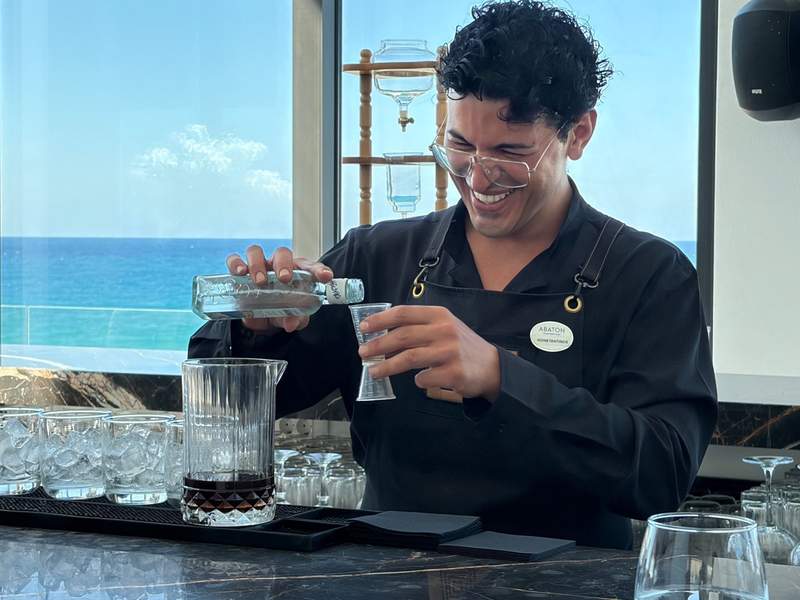 A smiling bartender mixes a custom cocktail. He has curly, dark hair and is wearing an all-black uniform. The blue Aegean Sea is visible behind him.