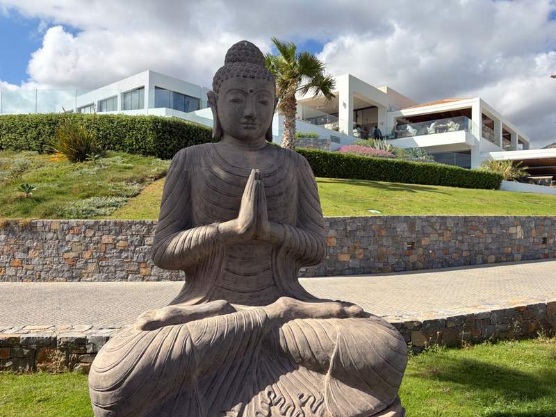 A stone statue of the Buddha in prayer is in the foreground, lush green space, stone walls and a white resort building with a pool area is in the background.