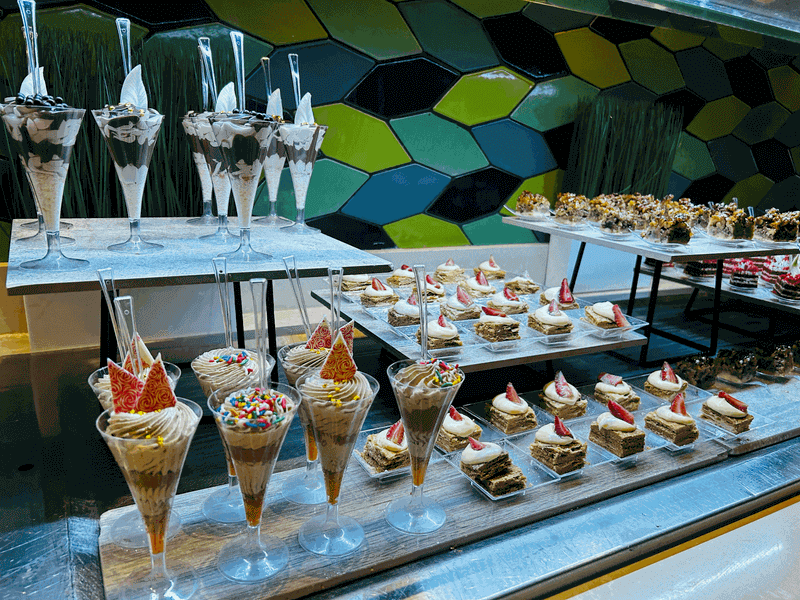 A vibrant close-up shot of rows of beautifully decorated, miniature pastries, colorful cakes, and parfaits lined up at a luxury resort buffet.