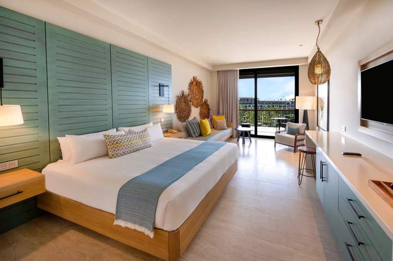 A bright, modern resort hotel room featuring a king-sized bed with crisp white linens, contemporary decor, and a view toward a tropical terrace.