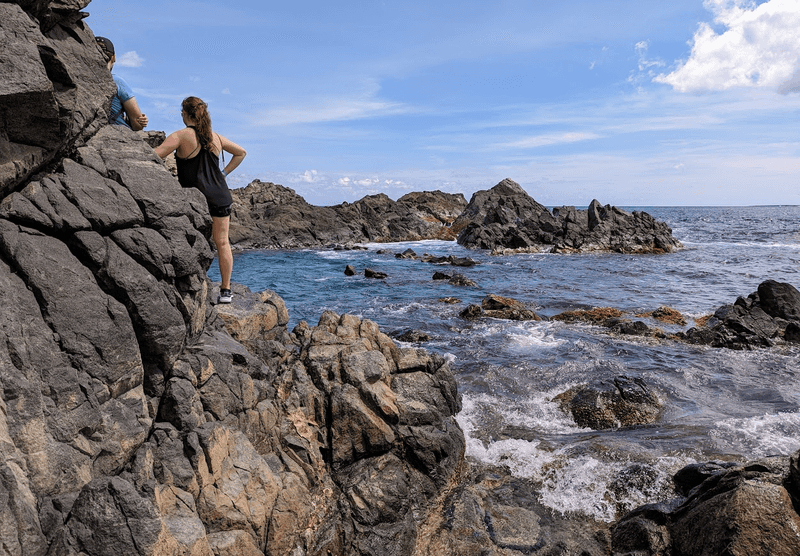 A woman standing on rugged coastal rocks next to a natural tide pool in Aruba.