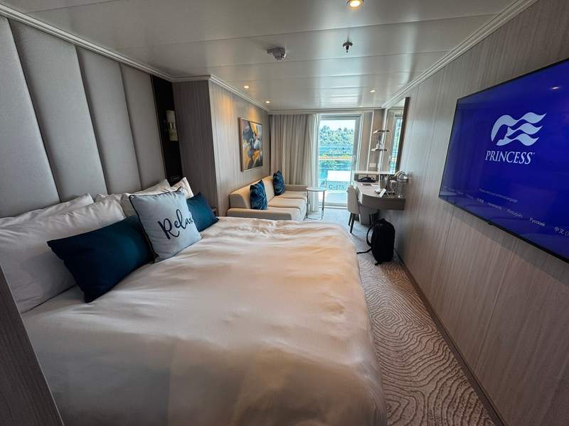 Deluxe balcony stateroom on Star Princess with queen bed, flat-screen TV, desk, sofa, and table and floor to ceiling sliding glass door.