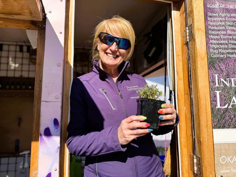 Woman standing in a doorway wearing a purple zip jacket with brightly colored nails, holding a lavender plant.