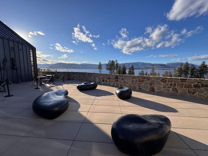 Black stones used as seating on a patio overlooking a lake. The patio has a stone wall, and you can see hills on the other side of the lake.