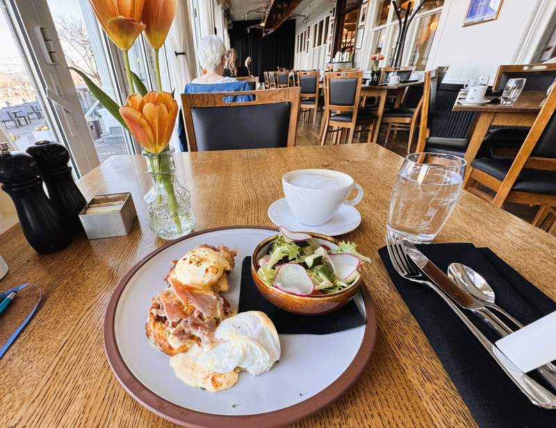 Eggs Benedict with a salad on a plate on top of a wooden table. Vase with flowers, coffee and a glass of water.