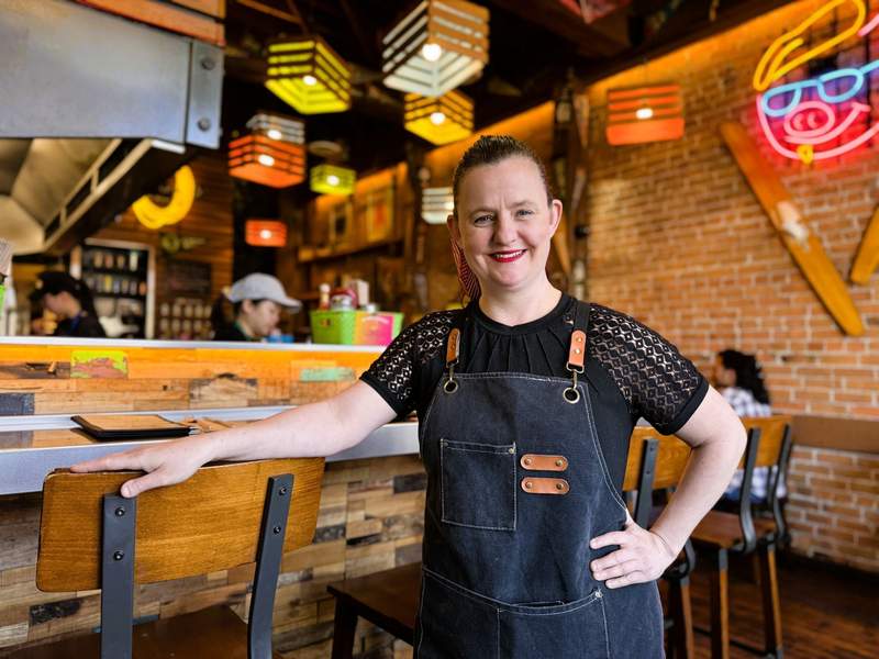 Chef Robyn Sigurdson wearing a denim apron, standing next to the bar with a brick wall behind her.