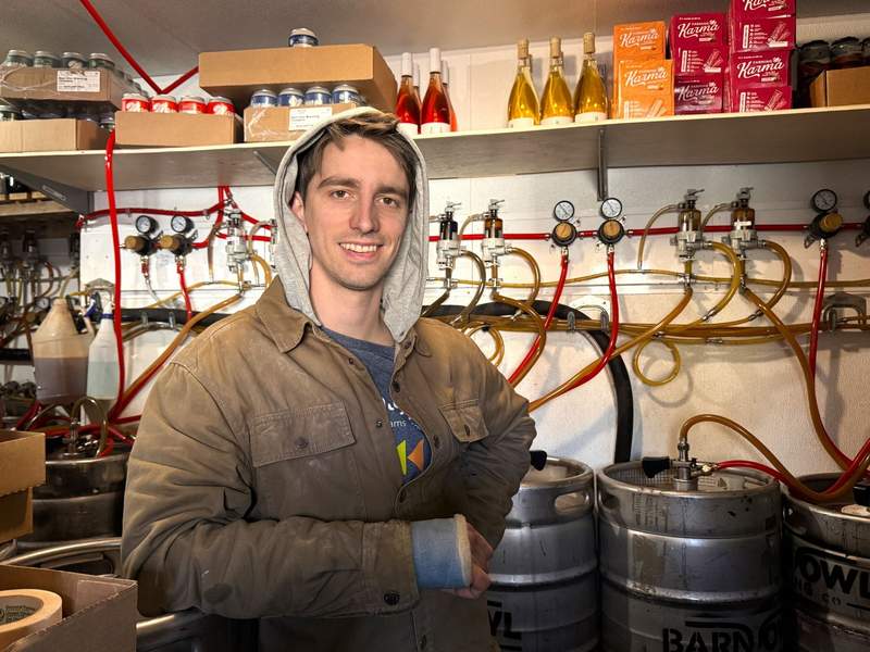 Colin Pistawka stands in front of barrels in the refrigerator at Barn Owl Brewing. He wears a warm jacket.
