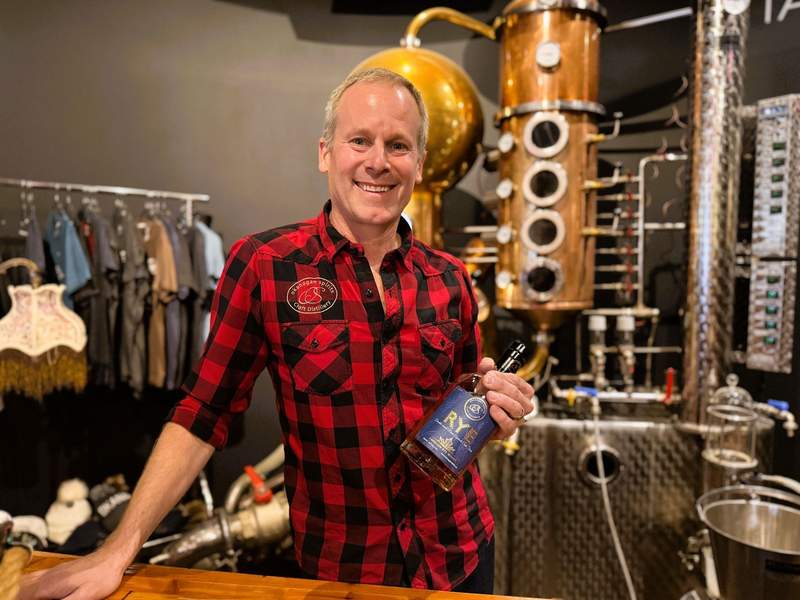 CEO Tyler Dyck wears a red-and-black checkered shirt while standing in front of a copper still, holding a bottle of Rye.