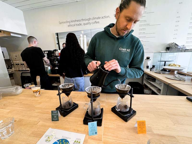 A man in a Craft 42 Roasters hoodie pours hot water over three pour-over coffees.