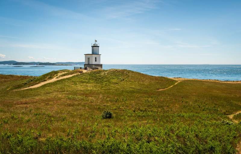 A small white lighthouse sitting on a windswept grassy hill overlooking the sea