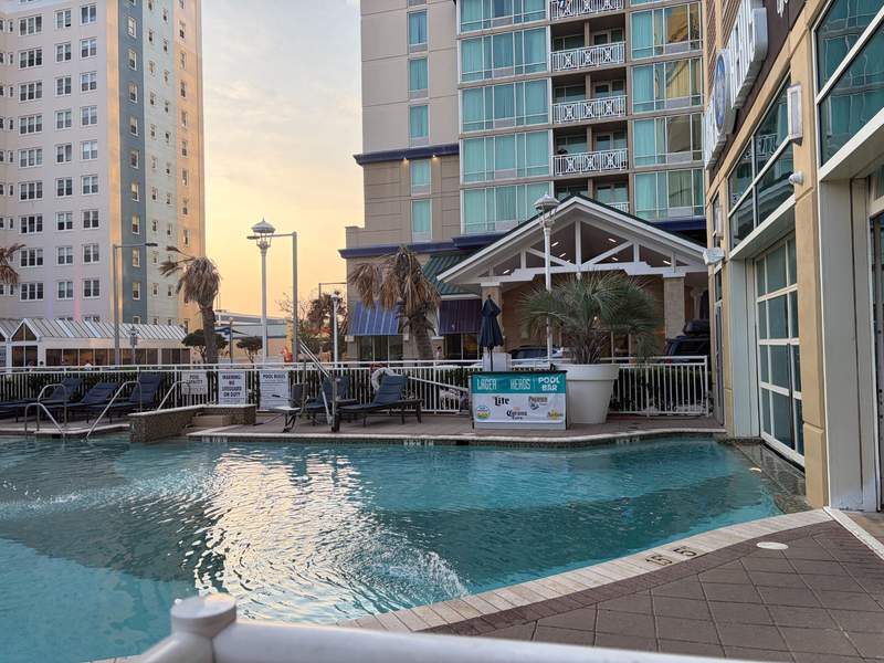 An outdoor resort-style swimming pool situated between two modern, high-rise beachfront hotel towers at sunset. The warm, golden evening light reflects off the water, creating a relaxing ambiance.