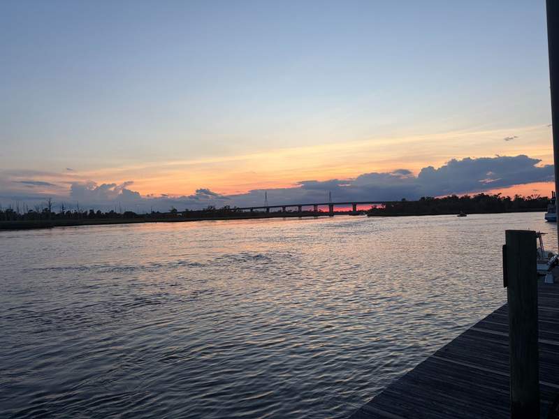 A tranquil coastal waterway at sunset along the Wilmington Riverwalk. Soft shades of pink, orange, and blue fill the evening sky, with a distant bridge reflecting on the calm water.