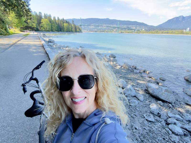 Kim Orlando with bike on the seawall trail around Stanley Park next to the Burrard Inlet and Lion's Gate Bridge in the backgroumd.