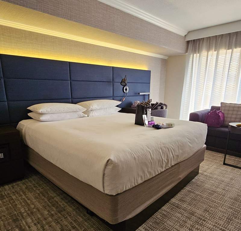 A bright hotel room featuring a plush queen bed with white linens, modern minimalist decor, and natural light from a window.