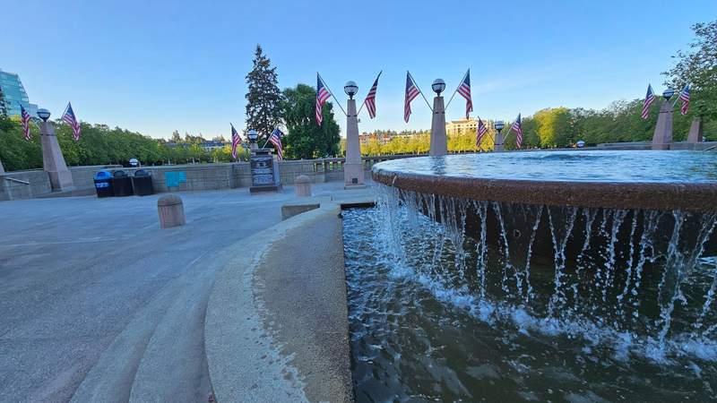 A wide paved path circling a green park lawn with a large modern water fountain at dawn.