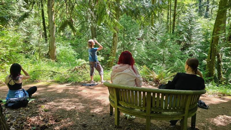 A small group of women standing in a lush green forest clearing surrounded by ferns and tall evergreen trees.