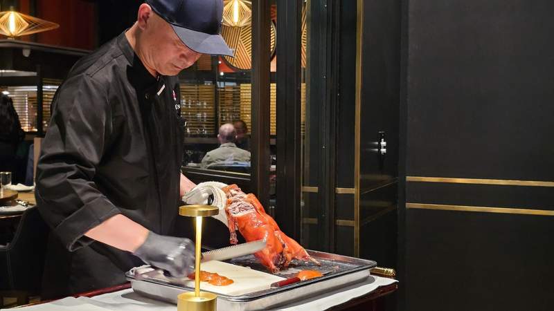 A chef carefully slicing a perfectly roasted, golden-brown whole Peking duck on a wooden carving board in a kitchen.