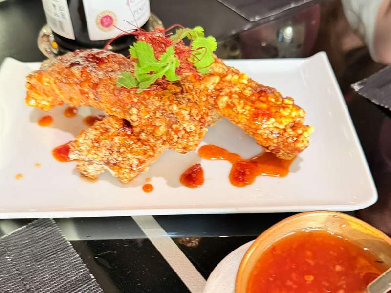Crunchy breaded ribs on a white plate with a small green cilantro sprig and red chili sauce in a bowl on the side.